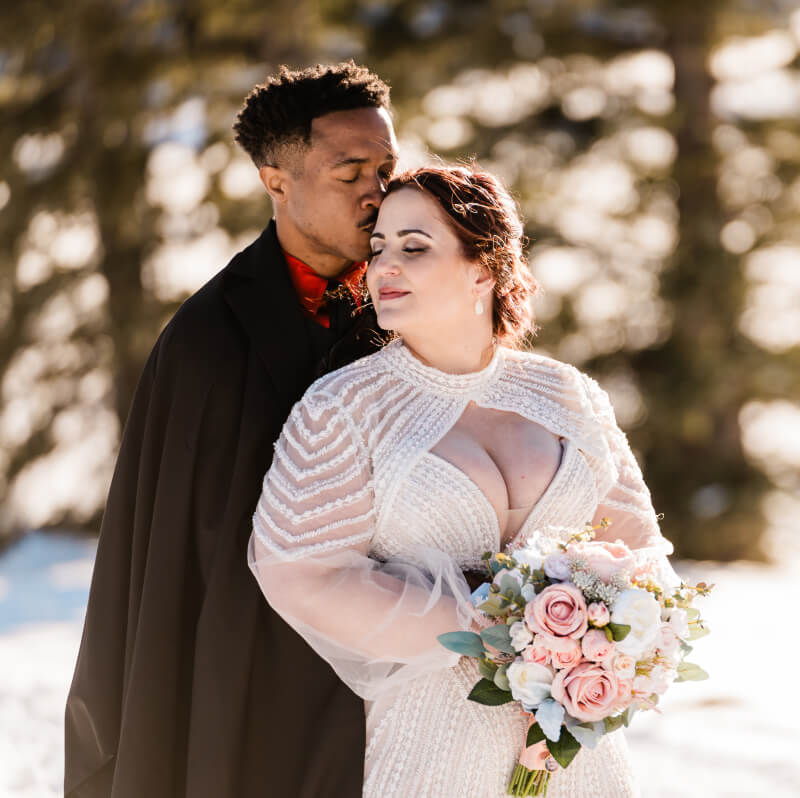 A person kisses their partner's forehead from behind, surrounded by snow.