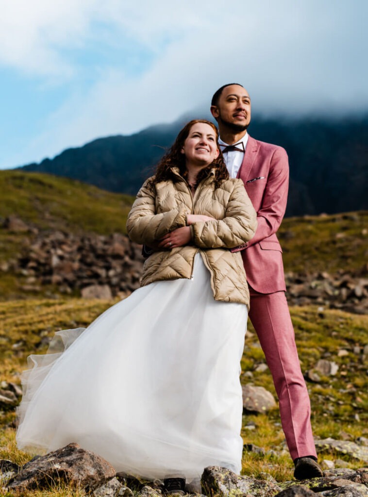 A couple holds one another, with smoke on green mountains behind them, as they look into the distance.
